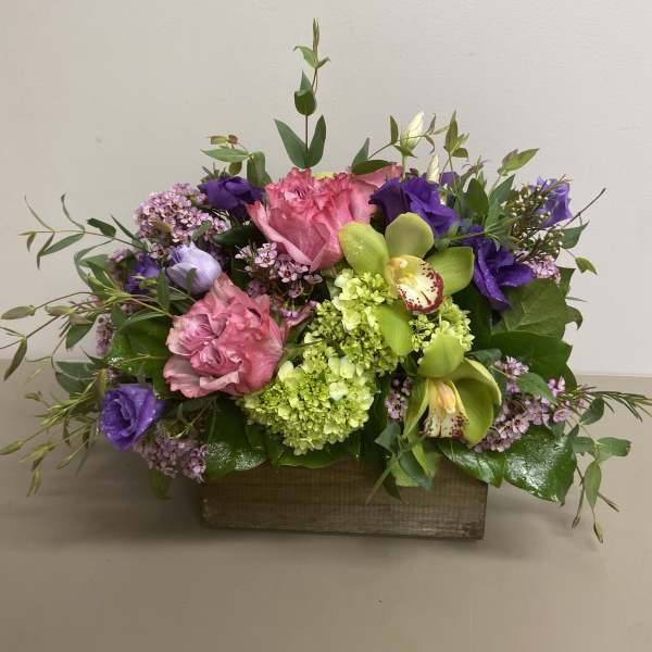 Low arrangement of pink roses, green hydrangeas, orchids and purple blooms in a rectangular wooden box