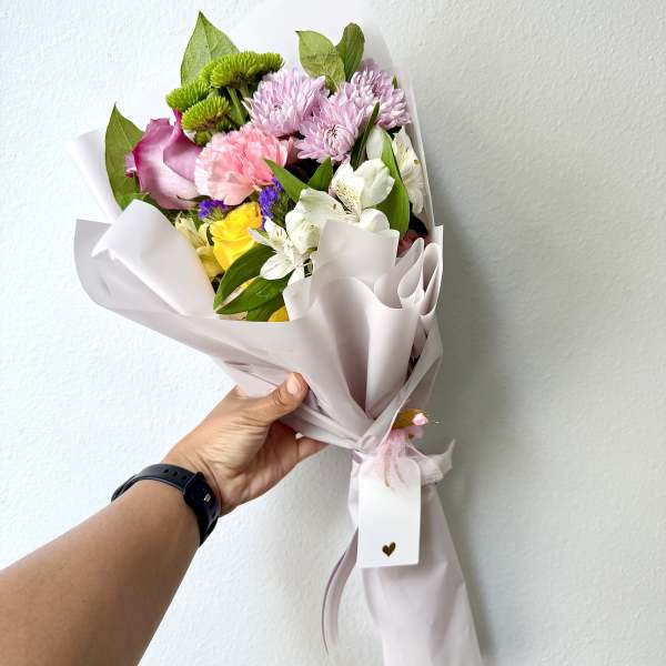 Handheld bouquet of pink, white, and yellow flowers wrapped in pale paper