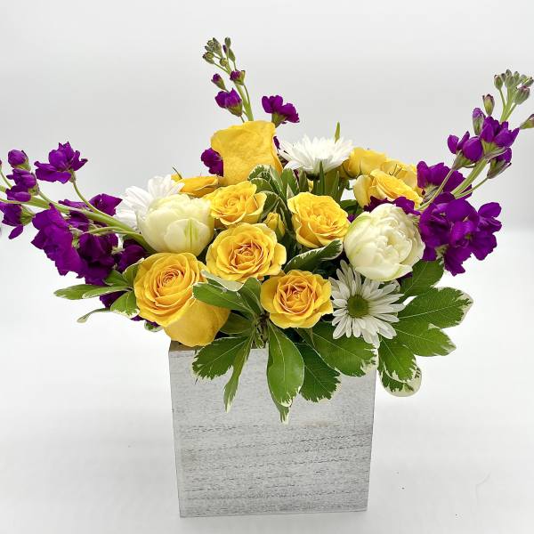 Yellow roses and white daisies in a square gray vase with purple flowers