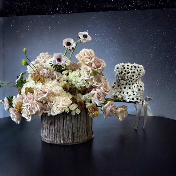 Blush roses and white anemones arranged in a rustic wood container beside a small white flower bear.
