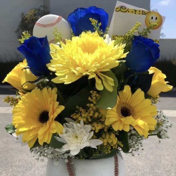 Yellow and blue flower arrangement with a baseball and smiley card in a white vase