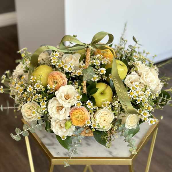 Basket of cream roses, ranunculus, and daisies with green apples and ribbon