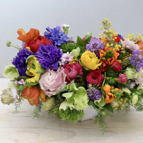 Low, oval arrangement of bright mixed flowers in a white ceramic container on a light wood surface
