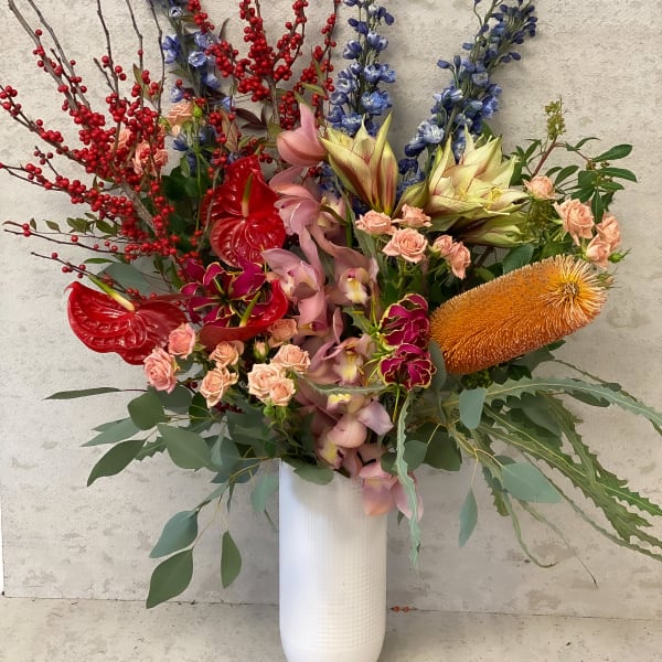 Tall mixed floral arrangement in a white vase with red berries and pink, blue, and orange blooms.