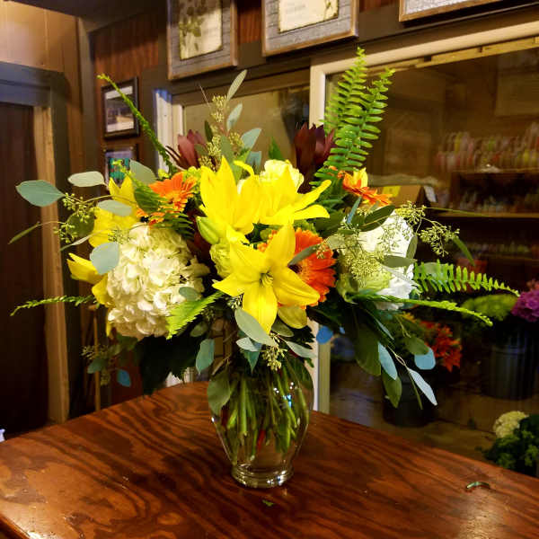 Mixed bouquet of yellow lilies, orange gerberas, white hydrangea, and greenery in a glass vase