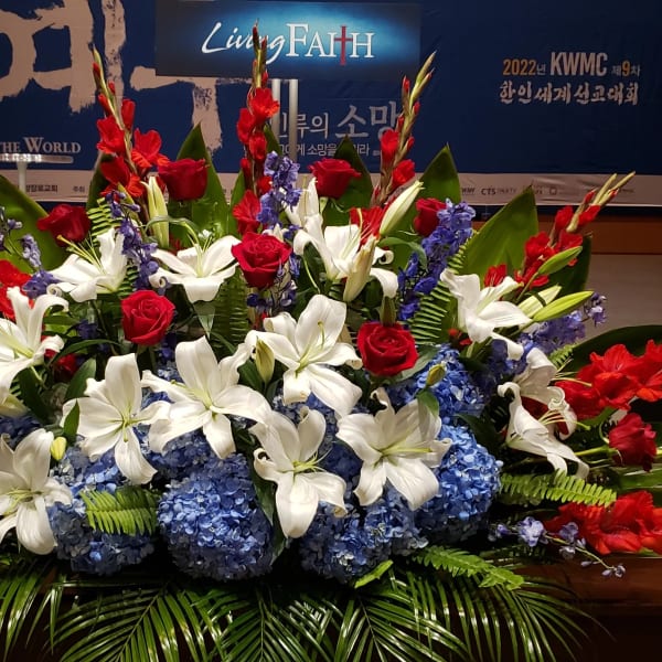 Large red, white, and blue stage arrangement with lilies, roses, hydrangeas, and palm fronds.