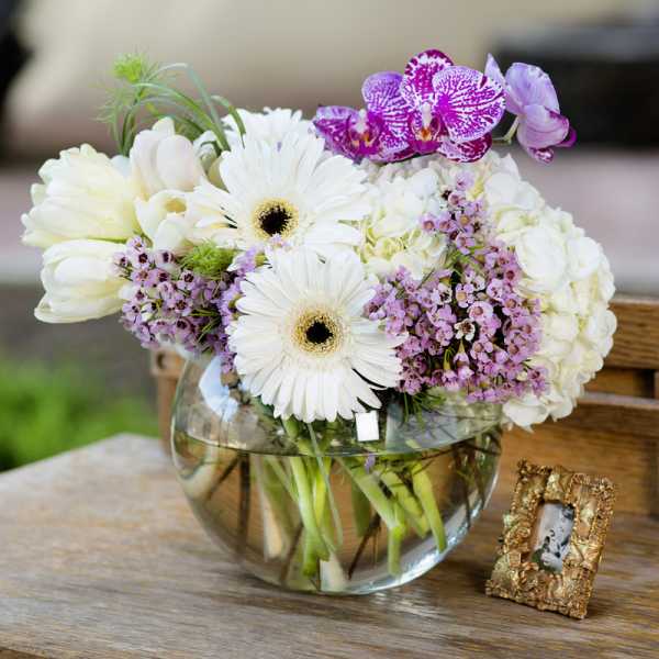 White and purple flowers arranged in a round glass vase on a table.