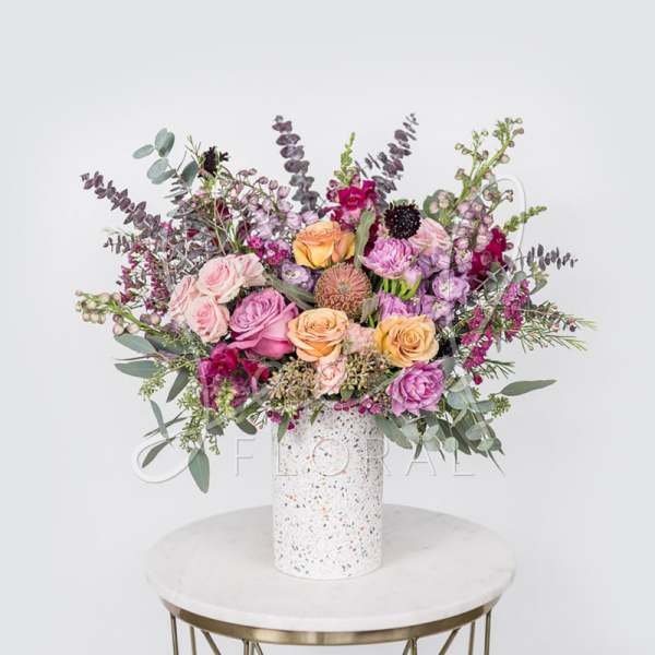 Mixed bouquet of pink, peach, and purple flowers in a speckled white vase