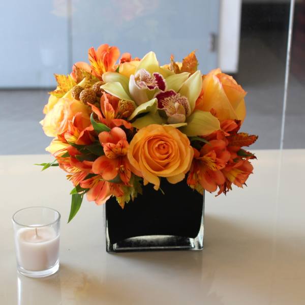 Orange and yellow floral arrangement in a square glass vase beside a small candle