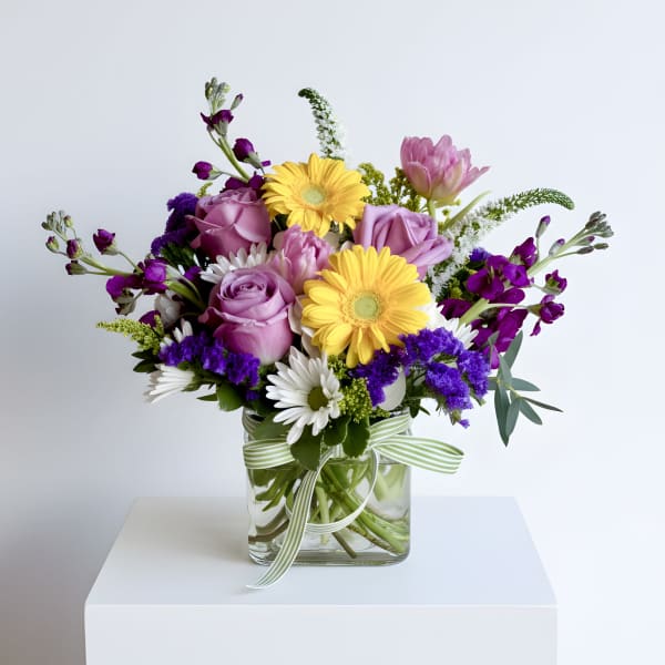 Mixed bouquet of purple roses, yellow gerberas and white daisies in a square glass vase with striped ribbon