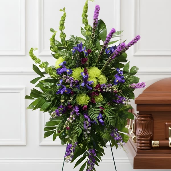 Tall standing spray of green mums, purple iris, and liatris on an easel beside a wooden casket
