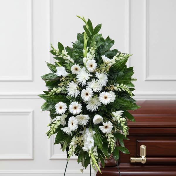 Standing spray of white gerbera daisies and mums with greenery on an easel beside a casket.