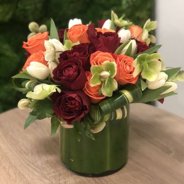 Compact arrangement of red and orange roses, white tulips, and green orchids in a leaf-wrapped cylinder vase