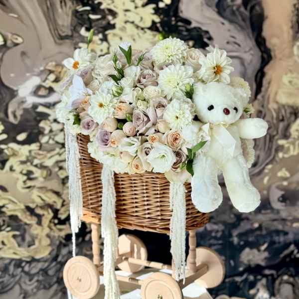 White floral basket arrangement with a teddy bear on a wooden cart
