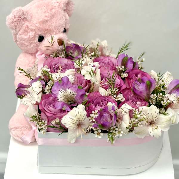 Pink teddy bear hugging a white hat box filled with pink and white flowers