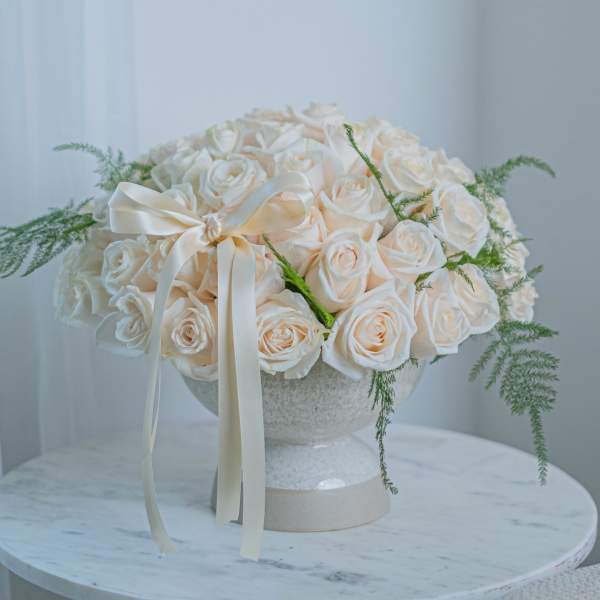 Cream rose bouquet in a white pedestal vase with a satin ribbon