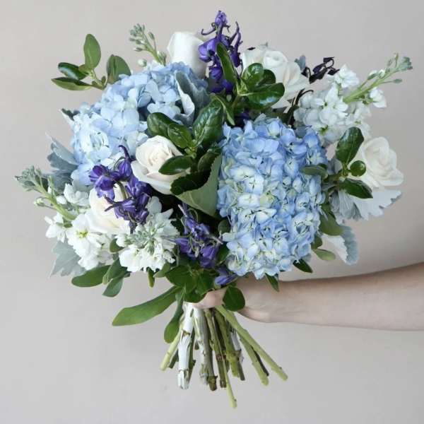 Handheld bouquet of blue hydrangeas, white roses, and purple flowers