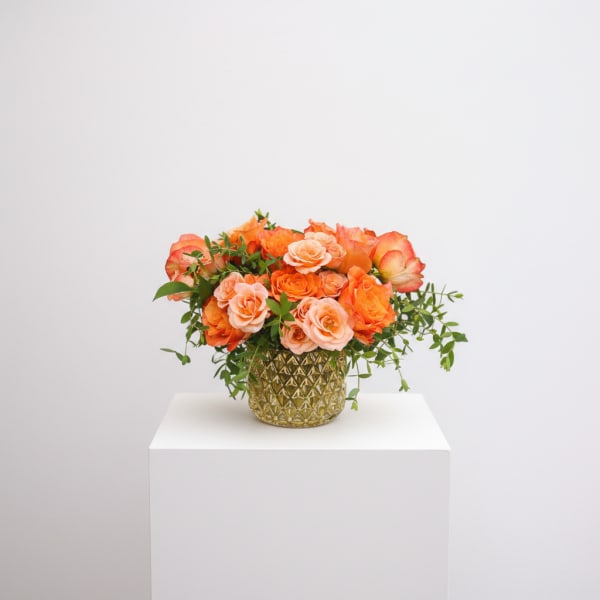 Low arrangement of orange and peach roses in a gold textured glass vase with trailing accents
