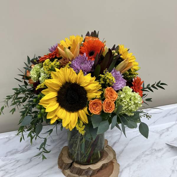 Mixed bouquet with sunflowers, orange roses, and purple blooms in a glass vase