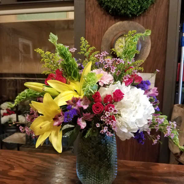 Mixed bouquet with yellow lilies, red roses, white hydrangea, and purple accents in a glass vase