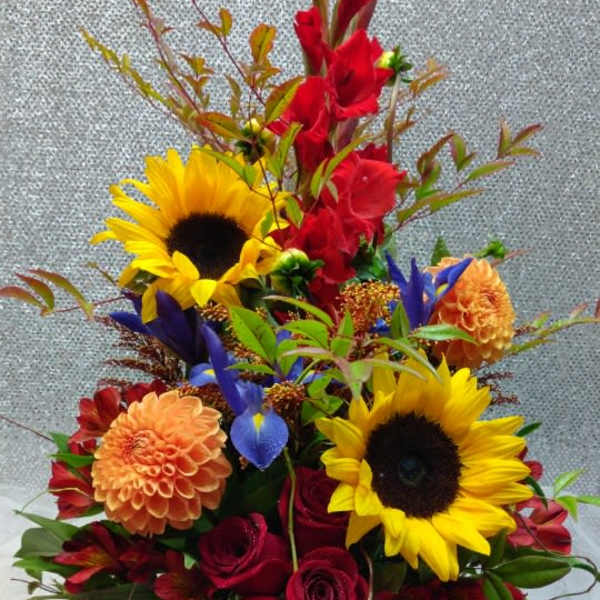 Tall mixed arrangement with sunflowers, red roses, orange dahlias, and purple iris in a clear glass vase