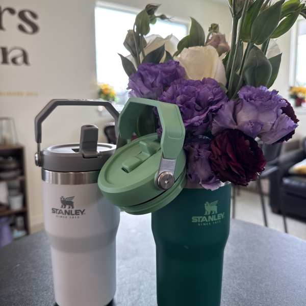 Purple and white flowers arranged in a green Stanley tumbler beside a white tumbler