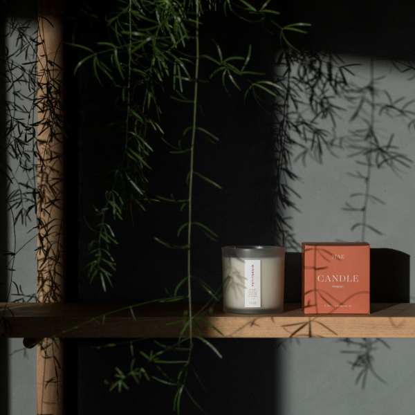 Candle in a glass jar beside its orange box on a wooden shelf