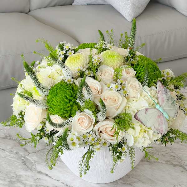 White and pale pink rose arrangement in a white bowl vase with green accents