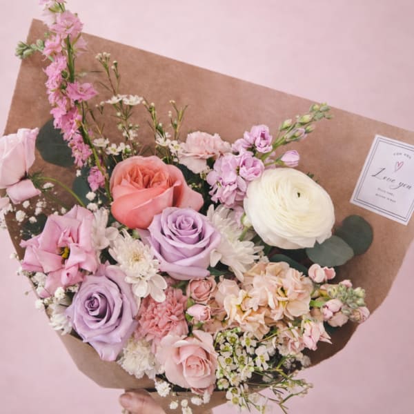 Handheld bouquet of pink, lavender, and white flowers wrapped in brown paper