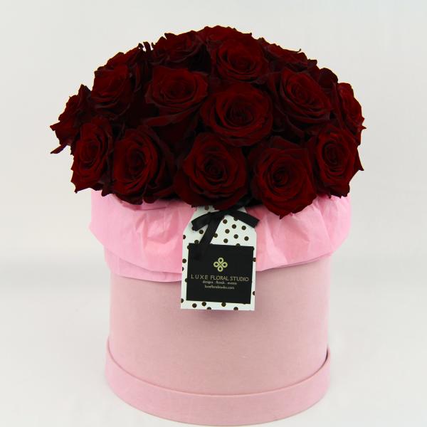 Red roses arranged in a pink round box with a hanging tag