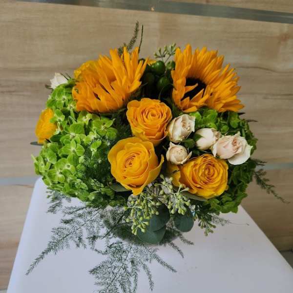 Round arrangement of yellow sunflowers and roses with green hydrangeas and cream spray roses.