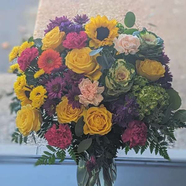 Bright mixed bouquet with yellow roses, sunflower, carnations, and mums in a clear glass vase