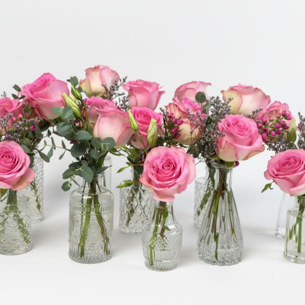 Multiple small glass bud vases holding pink roses and filler flowers against a white background