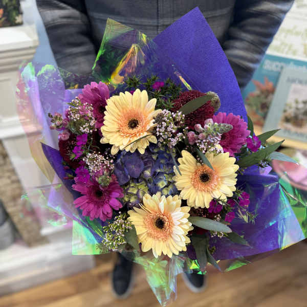 Hand-tied bouquet of yellow gerbera daisies and purple flowers wrapped in iridescent cellophane.