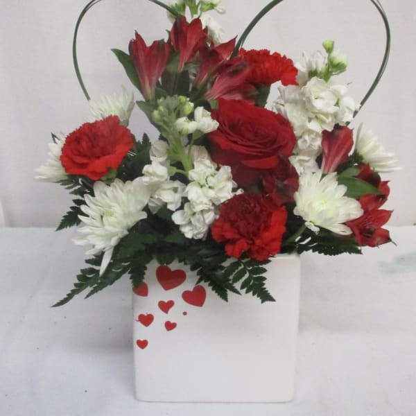 Red and white floral arrangement in a white heart-decorated container