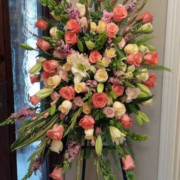 Tall standing spray of peach and cream roses with white lilies on a tripod easel.