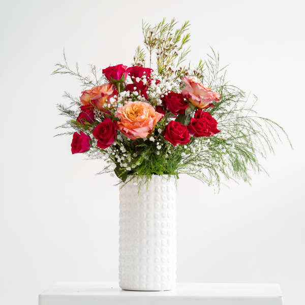 Tall arrangement of red and peach roses with white filler flowers in a textured white vase