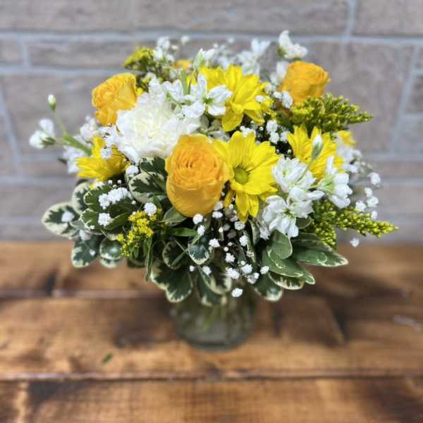 Yellow and white flower arrangement with roses and daisies in a clear glass vase on a wooden table.