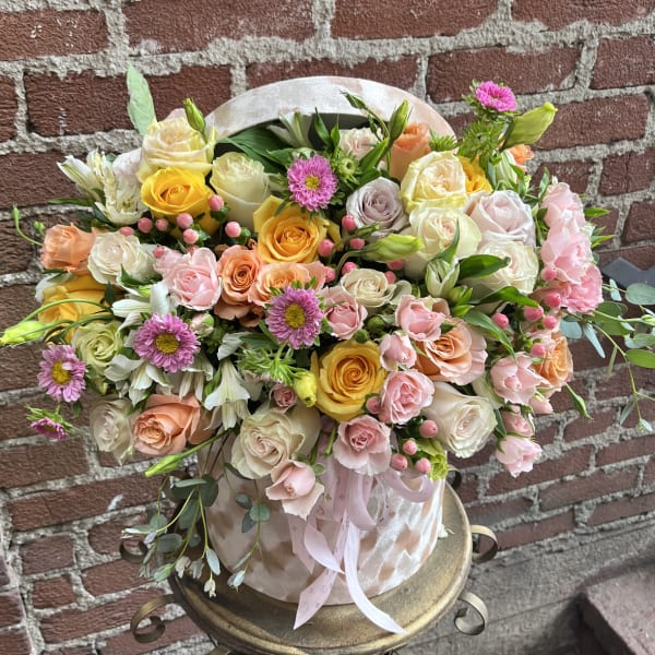 Pastel arrangement of roses, spray roses and daisylike blooms in a pink hatbox-style container