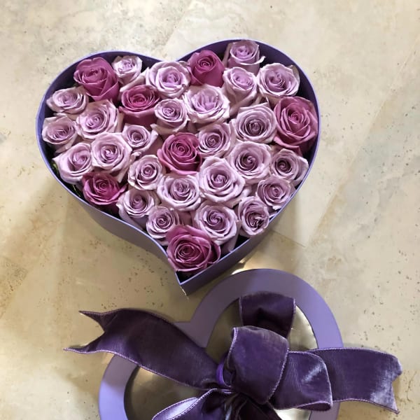 Heart-shaped box filled with pink and lavender roses with a purple ribbon bow