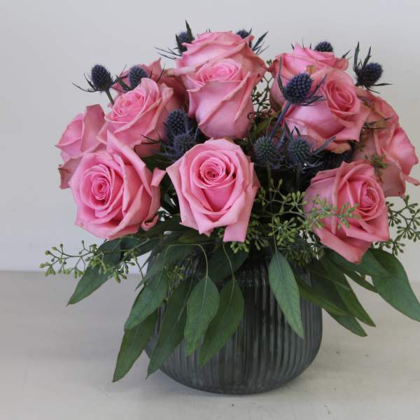 Pink roses arranged in a dark glass vase with blue thistle accents