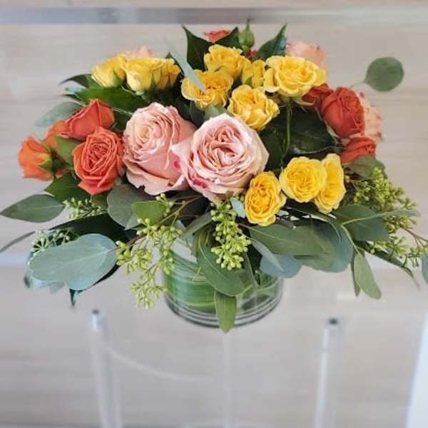 Round arrangement of yellow, peach, and orange roses in a clear glass cylinder vase