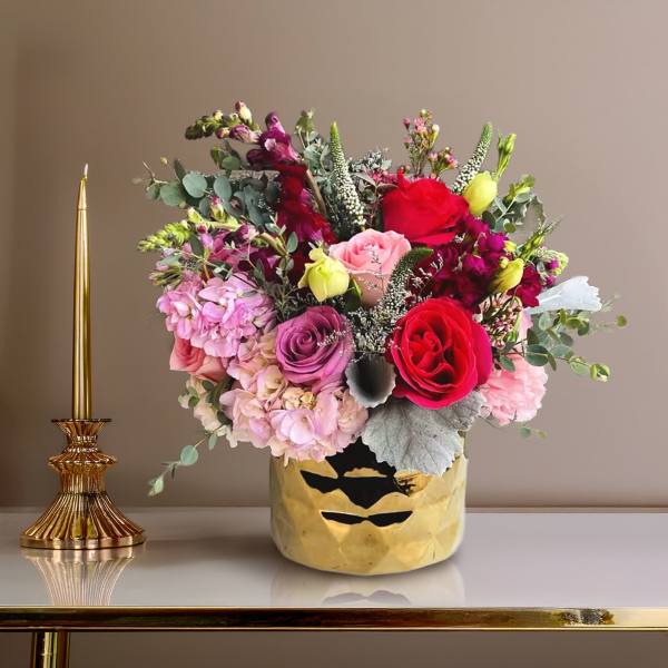 Mixed bouquet of pink and red flowers in a gold vase beside a gold candle holder