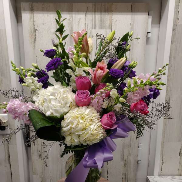 Tall mixed bouquet of pink, white, and purple flowers in a glass vase with a purple ribbon