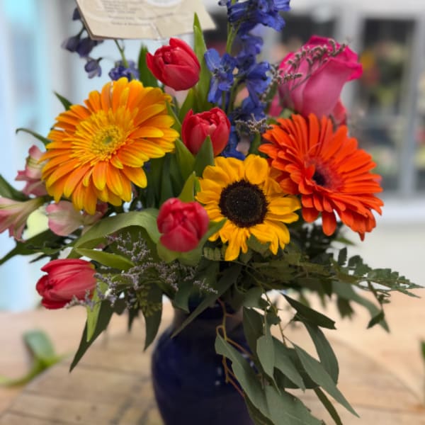 Bright mixed bouquet with orange gerbera daisies, a yellow sunflower, and pink tulips in a blue vase.
