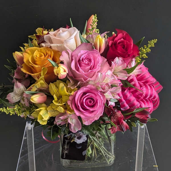 Compact arrangement of pink, yellow, and red roses with alstroemeria in a square glass vase with pink ribbon.