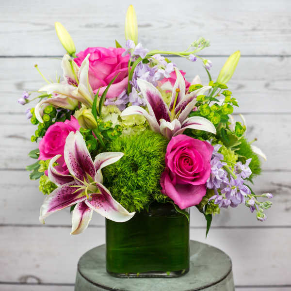 Bright arrangement of pink roses, white and magenta lilies, and lavender blooms in a green glass cube vase