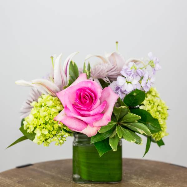 Pink rose arrangement in a green glass vase with pale purple and green flowers