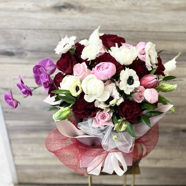 Round bouquet of red, pink, and white blooms with purple orchids and decorative red and white wrapping.