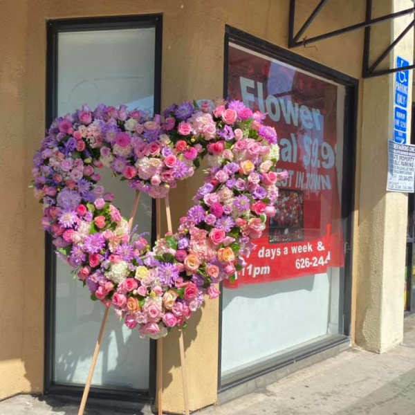 Large heart-shaped standing wreath of pink and lavender flowers on a wooden easel outside a flower shop